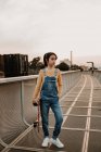 Стильна дівчина-підліток в джинсах в цілому з рукою в кишені тримає скейтборд на металевому мосту в місті — стокове фото