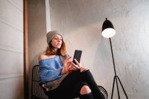 Молодая женщина в вязаном свитере с шарфом и шляпой на мобильном телефоне и сидя на стуле возле стены и лампы в комнате — стоковое фото