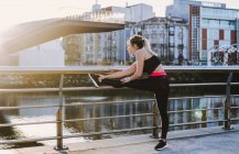 Женщина в спортивной одежде растягивает ноги на набережной возле воды в городе в солнечный день — стоковое фото
