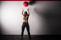 Спортивная молодая сконцентрированная леди в спортивной одежде в меле поднимает мячик тяжелой медицины на плечо в спортзале — стоковое фото