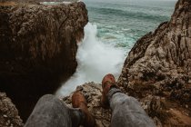 Обрезать ноги человека, сидящего на камне рядом с бурным морем в буфонах-де-приа, астурии, Испании — стоковое фото