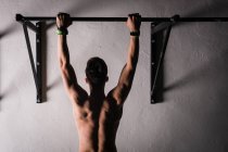 Rückenansicht des athletischen jungen Mannes ohne Hemd, der an der Stange neben der Wand in der Turnhalle hängt — Stockfoto