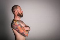 Боковой вид на бородатого хипстера со скрещенными руками и татуировками, смотрящего в сторону на сером фоне — стоковое фото