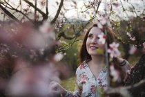 Vista a través de ramitas de árboles frutales florecientes de atractiva dama alegre mirando hacia el jardín - foto de stock