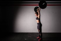 Vista laterale della giovane donna atletica concentrata in abbigliamento sportivo che alza il bilanciere sopra la testa in palestra — Foto stock