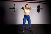 Атлетичная молодая сконцентрированная леди в спортивной одежде, поднимающая штангу над головой в спортзале — стоковое фото