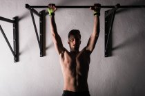 Athletischer junger Mann ohne Hemd hängt in Turnhalle an Stange neben Wand — Stockfoto