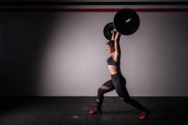 Вид сбоку на атлетичную молодую концентрированную леди в спортивной одежде, поднимающую штангу над головой в спортзале — стоковое фото