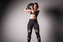 Atletica giovane donna concentrata in abbigliamento sportivo in gesso alzando pesante palla medica sulla spalla in palestra — Foto stock