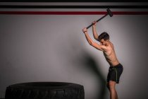 Shirtless guy concentrated in sportswear with hammer hitting on big tire in gym — Stock Photo