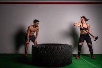 Homme torse nu debout près d'athlétique jeune femme concentrée en vêtements de sport avec marteau frapper sur un gros pneu dans la salle de gym — Photo de stock