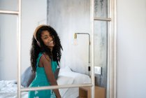 Black teenage girl with curly hair smiling looking at camera in front of a mirror listening to music in headphones — Stock Photo