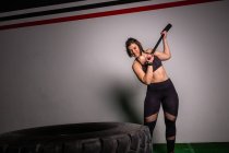 Атлетичная молодая сконцентрированная леди в спортивной одежде с молотком, ударяющим по большой шине в спортзале — стоковое фото