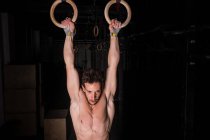 Athletic young shirtless guy hanging on gymnastic rings between obscurity in gym — Stock Photo