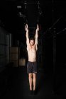Athletic young shirtless guy hanging on gymnastic rings between obscurity in gym — Stock Photo