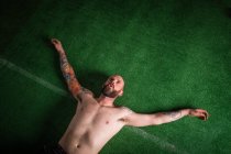 Dall'alto hipster stanco barbuto senza maglietta con tatuaggi e mani ai lati sdraiato sul tappeto erboso sintetico verde — Foto stock
