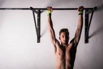 Athletic young shirtless guy hanging on bar near wall in gym — Stock Photo