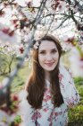 Vista attraverso ramoscelli di albero da frutto in fiore di attraente signora allegra guardando la fotocamera in giardino — Foto stock