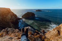 Crop legs of human sitting on top of stone hill near pitoresco sea in sunny day in Cabo de Penas, Astúrias, Espanha — Fotografia de Stock