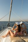 Вид сбоку на красивых молодых женщин в солнцезащитных очках и капитанских шляпах, сидящих на палубе дорогой лодки, плавающей на воде в солнечный день — стоковое фото