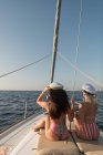 Вид сбоку на красивых молодых женщин в солнцезащитных очках и капитанских шляпах, сидящих на палубе дорогой лодки, плавающей на воде в солнечный день — стоковое фото