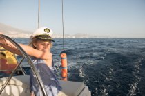 Positives Kind mit Kapitänsmütze schwimmt bei sonnigem Tag auf teurem Boot auf See — Stockfoto