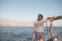 Позитивний бородатий дорослий чоловік в сонцезахисних окулярах плаває на дорогому човні на морі біля порту в сонячний день — стокове фото