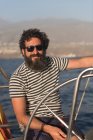 Positive bearded adult male in sunglasses floating on expensive boat on sea near port in sunny day — Stock Photo