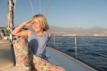Niño positivo con la mano cerca de la cara espiando desde el sol y sentado en la cubierta lateral de barco caro flotando en el mar - foto de stock