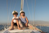 Позитивні діти в капелюхах капітана сидять на палубі дорогого човна, що плаває на воді в сонячний день — стокове фото