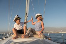 Позитивные дети в капитанских шляпах сидят на палубе дорогой лодки, плавающей по воде в солнечный день — стоковое фото