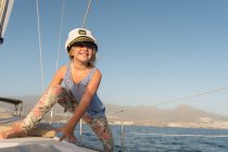 Позитивная девушка в капитанской шляпе сидит на палубе дорогой лодки, плавающей по воде в солнечный день — стоковое фото