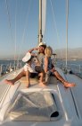 Positivo padre abbracciare i bambini felici in cappelli capitano e seduto sul ponte di barca costosa galleggiante sull'acqua nella giornata di sole — Foto stock