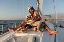 Позитивный отец, обнимающий счастливых детей в капитанских шляпах и сидящий на палубе дорогой лодки, плывущей по воде в солнечный день — стоковое фото