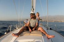 Позитивный отец с счастливыми детьми в капитанских шляпах и сидя на палубе дорогой лодки, плавающей на воде в солнечный день — стоковое фото