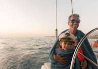 Positive father in sunglasses and towel embracing happy kid in captain hat and sitting on expensive boat floating on water — Stock Photo