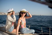 Vista lateral de belas fêmeas jovens em óculos de sol e chapéus de capitão no convés lateral do barco caro flutuando na água no dia ensolarado — Fotografia de Stock