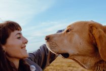 Side view of young hipster stroking funny dog between meadow and blue sky — Stock Photo