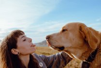 Side view of young hipster stroking funny dog between meadow and blue sky — Stock Photo