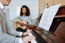 Вид сбоку на молодого человека, играющего на фортепиано рядом с черной женщиной, играющей на гитаре в музыкальной студии — стоковое фото