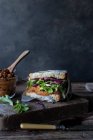 Sandwich of pate of dry tomatoes, fresh salad and cabbage on tray near knife on wooden board on black background — Stock Photo