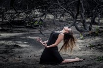 Молодая балерина в черной одежде позирует на земле между сухим лесом — стоковое фото