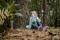 Позитивная маленькая девочка сидит рядом с деревом в лесу на размытом фоне — стоковое фото