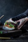 Couper les mains de la personne tenant le sandwich du pâté des tomates, la salade fraîche et le chou sur le plateau près du couteau sur la planche en bois — Photo de stock