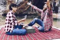 Вид сбоку мужчины и женщины в клетчатых рубашках, играющих в карты на клетчатых пикниках на берегу озера в скалах — стоковое фото