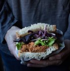 Руки человека, держащего сэндвич с помидорами, свежий салат и капуста на подносе рядом с ножом на деревянной доске — стоковое фото