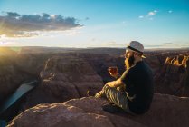 Бородатый парень наслаждается горячим напитком и смотрит на закат солнца, сидя рядом с прекрасным каньоном на западном побережье США — стоковое фото