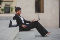 Очаровательная уверенная афро-американская элегантная женщина в костюмах просматривает ноутбук и сидит на лестнице рядом с сумкой на улице — стоковое фото
