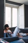 Attraktive junge stylische Frau mit Mütze trinkt Kaffee und ruht sich zu Hause auf dem Sofa in der Nähe des Balkons aus — Stockfoto