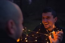 Closeup of cheerful men untangling illuminated fairy lights in murk forest in evening on blurred background — Stock Photo